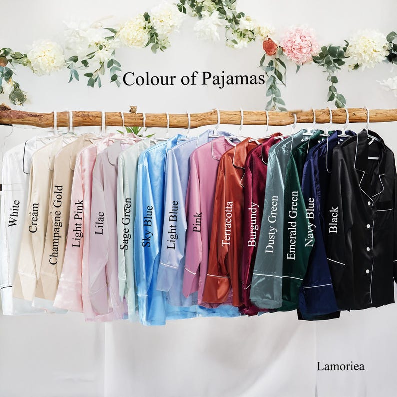 Puede incluir: Una colecci&oacute;n de pijamas de sat&eacute;n en varios colores, incluyendo blanco, crema, rosa, azul y negro, colgados de una barra de madera. Cada conjunto de pijama tiene una camisa con botones y se muestra con una etiqueta blanca que indica el color.