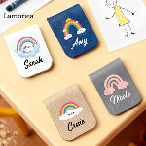 Puede incluir: Una colecci&oacute;n de marcadores personalizados de polipiel en varios colores, cada uno con un dise&ntilde;o de arco&iacute;ris y un nombre. Los marcadores son blancos, azul marino, beige y gris. Los nombres "Sarah", "Amy", "Cassie" y "Nicole" est&aacute;n impresos.