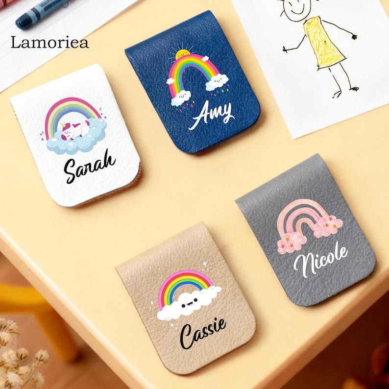 Puede incluir: Una colecci&oacute;n de marcadores personalizados de polipiel en varios colores, cada uno con un dise&ntilde;o de arco&iacute;ris y un nombre. Los marcadores son blancos, azul marino, beige y gris. Los nombres "Sarah", "Amy", "Cassie" y "Nicole" est&aacute;n impresos.
