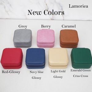 May include: A selection of square jewelry cases in various colors, including grey, berry, caramel, red-glossy, navy blue, light gold, and emerald green. Each case has a zipper closure and is labeled with its color name. The text "New Colors" is displayed at the top.