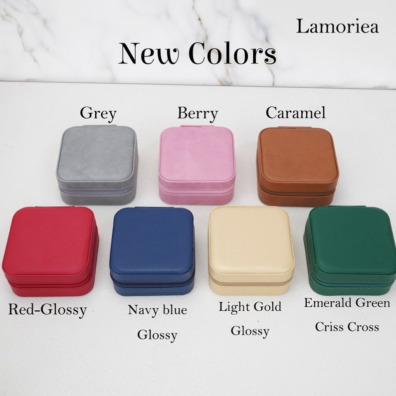 May include: A selection of square jewelry cases in various colors, including grey, berry, caramel, red-glossy, navy blue, light gold, and emerald green. Each case has a zipper closure and is labeled with its color name. The text "New Colors" is displayed at the top.