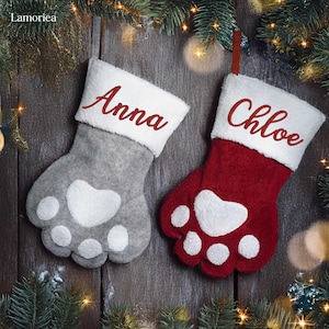 May include: Two personalized Christmas stockings with paw print designs. One stocking is gray with the name "Anna" embroidered in red. The other stocking is red with the name "Chloe" embroidered in white.