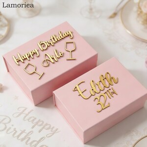 May include: Two rectangular light pink gift boxes with gold-coloured lettering. One box features the text Happy Birthday Arlo with two wine glass icons. The second box displays the name Edith and the text 32TH in a cursive gold-coloured font.