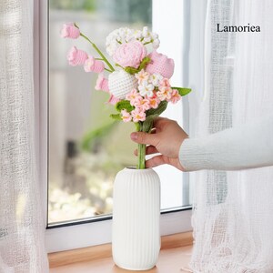 May include: A bouquet of crocheted flowers in pink and white, arranged in a white vase. The flowers are made of yarn and have a realistic appearance. The vase is tall and slender, with a ribbed texture. The image is captioned "Lamorie