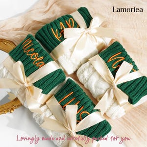 May include: Four personalized knitted gifts, each wrapped with a cream-colored ribbon. The gifts feature green and white sections, with embroidered names in orange. The text "Lamoriea" and "Lovingly made and carefully packed for you" are also visible.