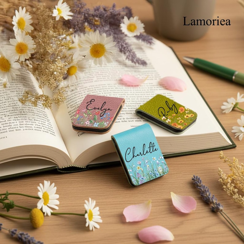 May include: Three personalized magnetic bookmarks with floral designs and names: "Evelyn" on pink, "Ruby" on green, and "Charlotte" on blue. The bookmarks are displayed on an open book, surrounded by flowers and petals.