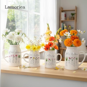Pode incluir: Quatro vasos de cerâmica branca com alças, cada um cheio de flores coloridas. Os vasos têm escrita preta e desenhos florais. O texto nos vasos diz "Oma's Garden", "Mr. Lindsey", "Mommy" e "Mr. Wilson".