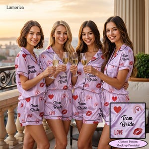 May include: Four women in matching light purple Team Bride pyjama sets, toasting with champagne. The sets have red hearts, lip prints, and rainbow designs. The text "Team Bride" and "LOVE" are printed on the pyjamas.