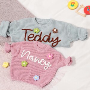 May include: A light blue knit jumper with the brown embroidered text Teddy and small green and brown flower appliqu&eacute;s, and a pink knit jumper with the white embroidered text Nancy and small green, pink, and blue flower appliqu&eacute;s.