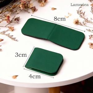 May include: Two green leather accessories, one rectangular (8cm long) and one square (4cm x 3cm), are displayed on a white surface. The accessories are likely small wallets or cardholders. The brand name "Lamoriea" is visible in the top right corner.
