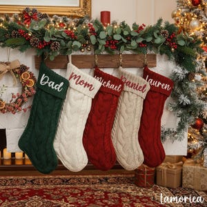 May include: Five cable-knit Christmas stockings hang above a fireplace. The stockings are green, cream, and red, each with a name embroidered in a contrasting color: "Dad", "Mom", "Michael", "Daniel", and "Lauren". A festive garland and Christmas tree are in the background.