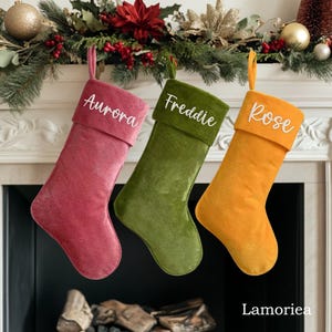 May include: Three velvet Christmas stockings with personalised names: Aurora, Freddie, and Rose. The stockings are hung on a fireplace mantel with a garland of greenery and red berries.