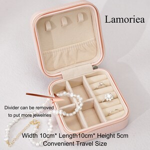 May include: A pink, zippered jewelry case with an open top, revealing compartments for rings and earrings. The case is labeled "Lamoriea" and measures 10cm x 10cm x 5cm. The text "Divider can be removed to put more jewelries" is also visible.