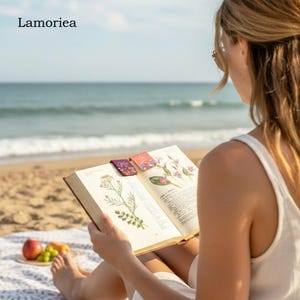 May include: A person reading a book on a beach with three decorative magnetic bookmarks. The bookmarks are rectangular with floral designs and the word "Me" on one. The book is open to a page with botanical illustrations. The word "Lamoriea" is in the top left corner.