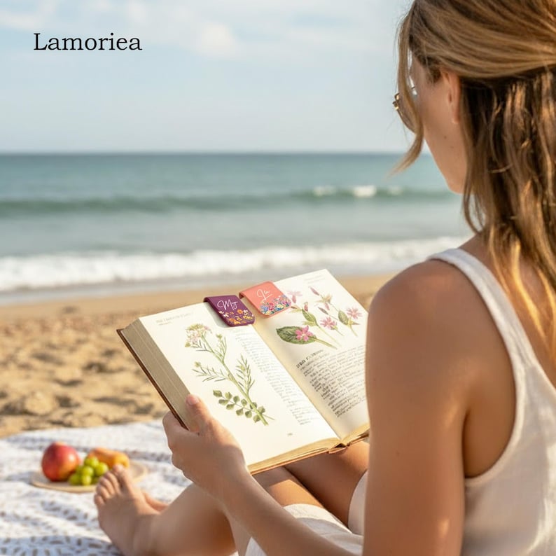 May include: A person reading a book on a beach with three decorative magnetic bookmarks. The bookmarks are rectangular with floral designs and the word "Me" on one. The book is open to a page with botanical illustrations. The word "Lamoriea" is in the top left corner.