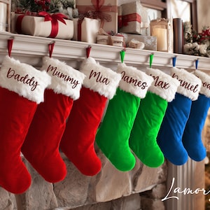 Op de afbeelding: Een rij gepersonaliseerde kerstsokken in rood, groen en blauw, hangend aan een schoorsteenmantel. Elke sok heeft een witte imitatiebont manchet en een naam geborduurd in een sierlijk lettertype. De namen zijn "Daddy", "Mummy", "Mia", "James", "Max", "Daniel" en "Rody".