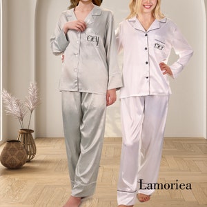 Puede incluir: Dos conjuntos de pijama de sat&eacute;n. Uno es gris claro con ribete blanco, el otro es blanco con ribete negro. Cada conjunto tiene una camisa abotonada con iniciales bordadas y pantalones a juego. La marca "Lamoriea" es visible.