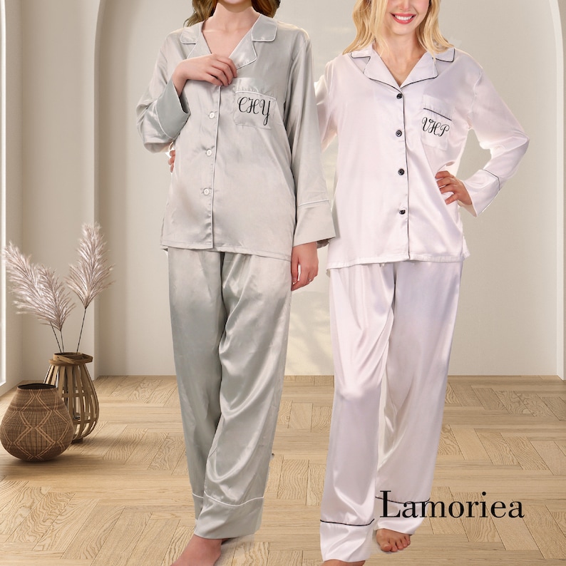 Puede incluir: Dos conjuntos de pijama de sat&eacute;n. Uno es gris claro con ribete blanco, el otro es blanco con ribete negro. Cada conjunto tiene una camisa abotonada con iniciales bordadas y pantalones a juego. La marca "Lamoriea" es visible.