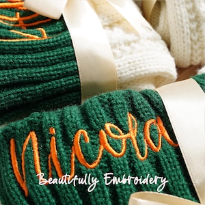 May include: Close-up of green knit items with orange embroidered text that reads "Nicola". The words "Beautifully Embroidery" are below the name. Cream-colored ribbon is tied around the items. White knit items are in the background.