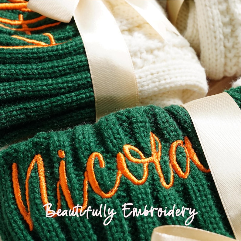 May include: Close-up of green knit items with orange embroidered text that reads "Nicola". The words "Beautifully Embroidery" are below the name. Cream-colored ribbon is tied around the items. White knit items are in the background.