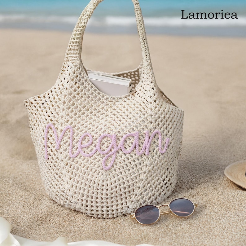 Peut inclure: Un sac fourre-tout en maille ajour&eacute;e de couleur cr&egrave;me avec le nom "Megan" brod&eacute; en lavande. Le sac a une anse et contient un livre. Des lunettes de soleil &agrave; monture dor&eacute;e reposent sur le sable pr&egrave;s du sac.
