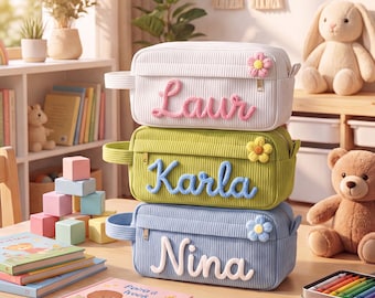 May include: Three rectangular corduroy pencil cases stacked vertically. The top case is white with pink text reading Laur and a pink flower. The middle case is olive green with blue text reading Karla and a yellow flower. The bottom case is light blue with white text reading Nina and a blue flower.