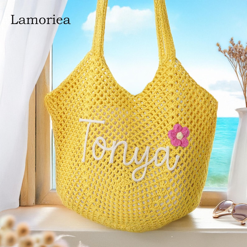 Peut inclure: Un sac fourre-tout jaune en crochet avec le nom "Tonya" brod&eacute; en blanc. Une petite fleur rose est attach&eacute;e au sac. Le sac a deux anses et est pos&eacute; sur un rebord de fen&ecirc;tre.