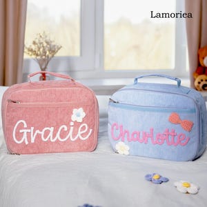 Op de afbeelding: Twee corduroy lunchtassen, één roze en één lichtblauw, elk met een naam op de voorkant geborduurd. De roze tas zegt "Gracie" en heeft een witte bloem. De blauwe tas zegt "Charlotte" en heeft een roze strik.