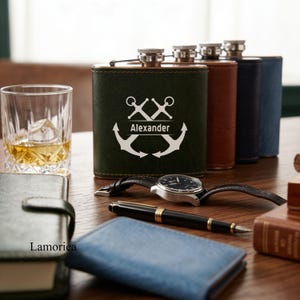 May include: A collection of personalized flasks in various colors, including green, brown, navy, and blue. The green flask features an anchor design with the name "Alexander." A watch, pen, and small leather goods are also on the wooden surface.