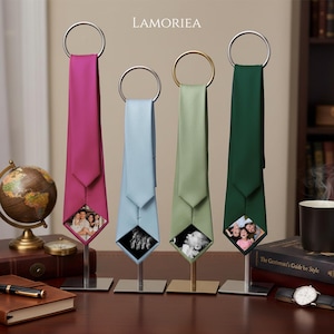 May include: Four neckties in magenta, light blue, sage green, and dark green, each hanging from a circular metal ring on a vertical stand. The bottom tip of each tie features a square photo insert displaying various family and portrait images.