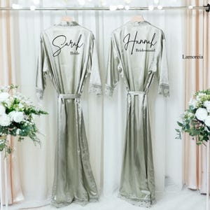Könnte beinhalten: Zwei silbergrüne Satinroben hängen an einer Stange. Eine Robe ist mit "Sarah Bride" in schwarzer Schrift beschriftet, die andere mit "Hannah Bridesmaid". Beide Roben haben Spitzentrimm an den Ärmeln und am Saum sowie passende Bindegürtel. Die Roben werden vor einem weißen Vorhang präsentiert.