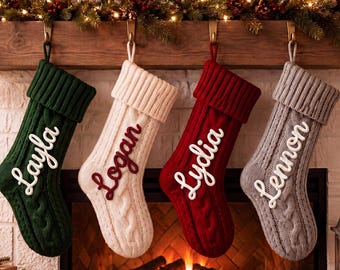May include: Four cable-knit Christmas stockings hanging in a row. The first is dark green with white text reading Layla, the second is cream with red text reading Logan, the third is red with white text reading Lydia, and the fourth is gray with white text reading Lennon.