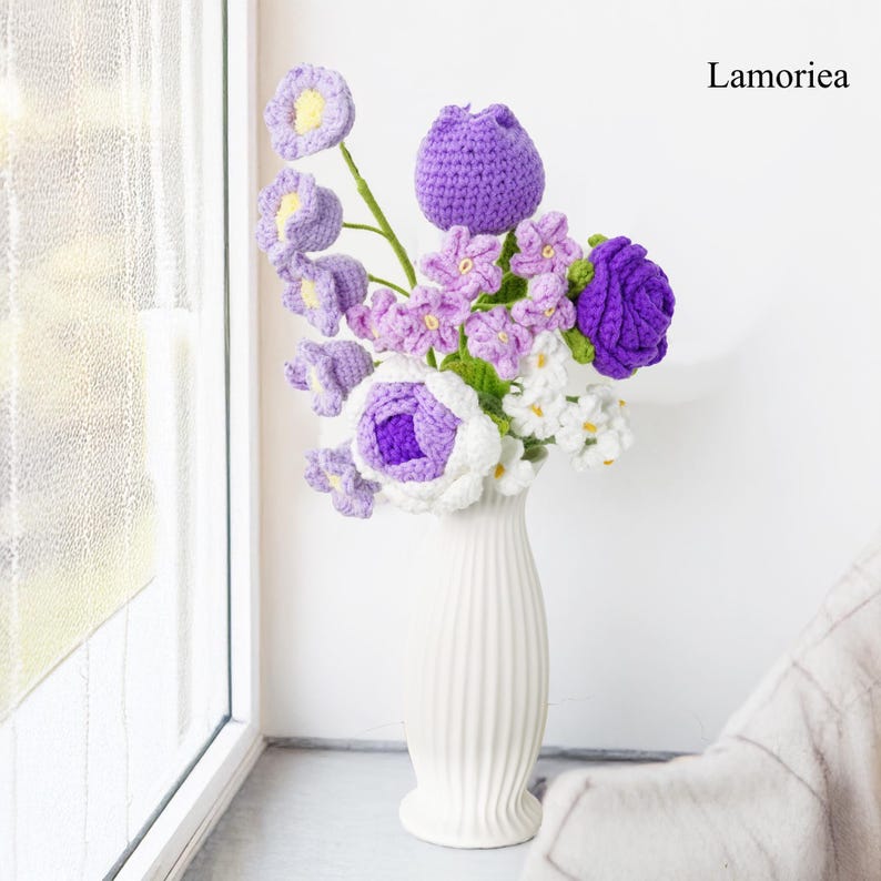 May include: A bouquet of crocheted flowers in shades of purple and white, arranged in a white vase. The flowers are made with yarn and have a realistic appearance. The text "Lamorie" is visible in the upper right corner of the image.
