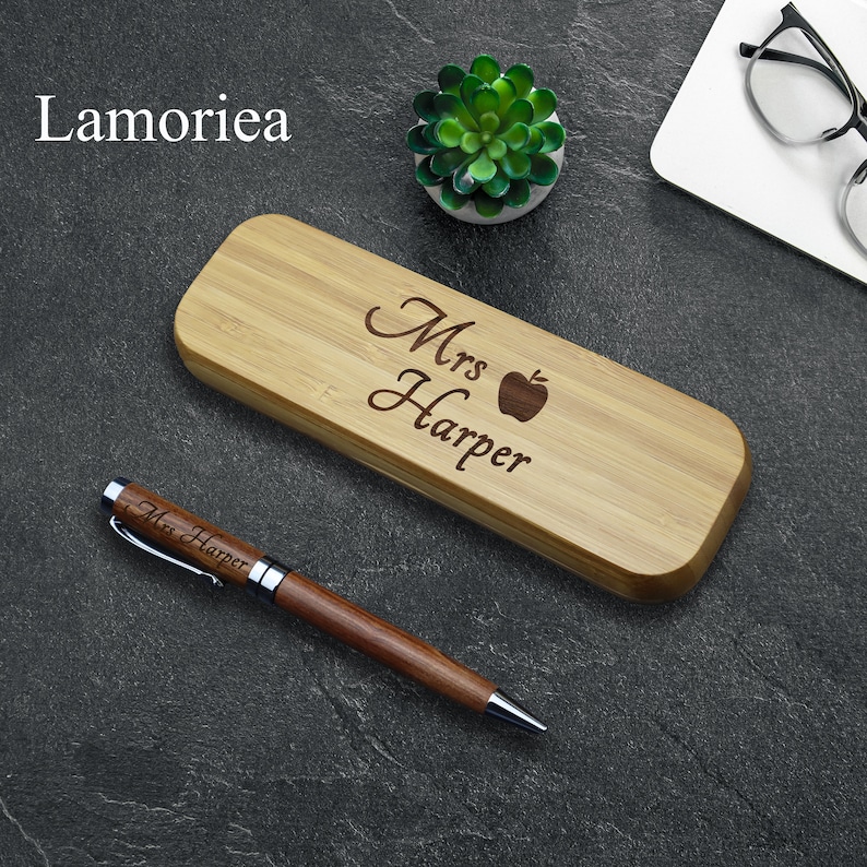 May include: A wooden pen and pencil holder with the text "Mrs. Harper" and an apple engraved on it. The holder is made of light brown bamboo and has a natural finish. A matching engraved pen is also included.