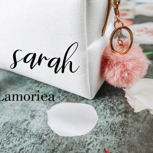 May include: White canvas makeup bag with the name "Sarah Lamorie" printed on it. The bag has a pink fluffy keychain attached to the zipper.