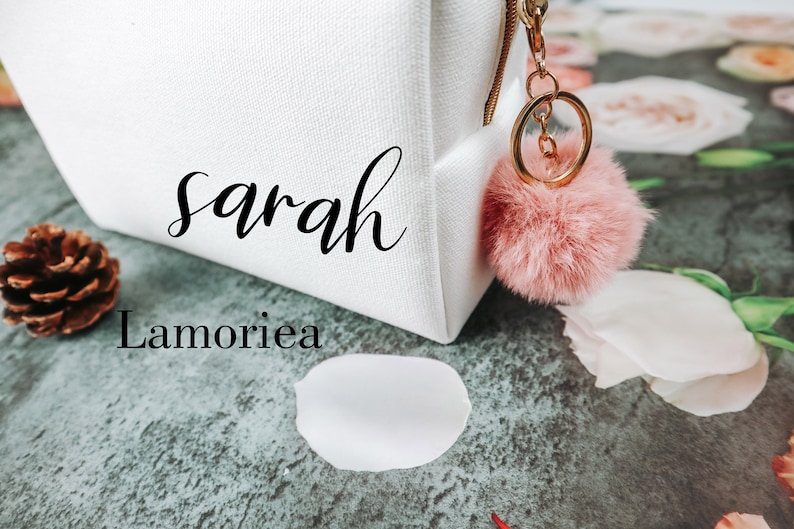 May include: White canvas makeup bag with the name "Sarah Lamorie" printed on it. The bag has a pink fluffy keychain attached to the zipper.