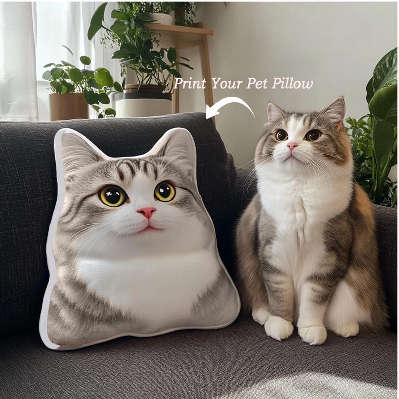 Puede incluir: Un coj&iacute;n con forma de gato y un gato real est&aacute;n sentados en un sof&aacute;. El coj&iacute;n tiene un patr&oacute;n atigrado blanco, gris y marr&oacute;n, con ojos amarillos y una nariz rosa. El texto "Print Your Pet Pillow" est&aacute; encima del coj&iacute;n.