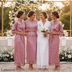 Op de afbeelding: Vier vrouwen in zijden robes, drie in oudroze en één in wit, staan samen. De oudroze robes hebben kanten randen en de initialen D, E en H. De witte robe heeft de initiaal K. Een tafel is gedekt met bloemen.