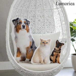 Puede incluir: Tres cojines con forma de mascotas: un pastor australiano, un gato color crema y un cachorro de pastor alem&aacute;n. Los cojines est&aacute;n sobre un coj&iacute;n blanco dentro de una silla colgante blanca tejida. La marca "Lamorea" es visible.