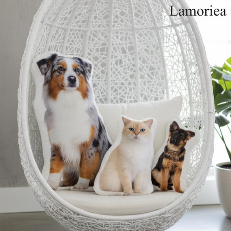 Puede incluir: Tres cojines con forma de mascotas: un pastor australiano, un gato color crema y un cachorro de pastor alem&aacute;n. Los cojines est&aacute;n sobre un coj&iacute;n blanco dentro de una silla colgante blanca tejida. La marca "Lamorea" es visible.