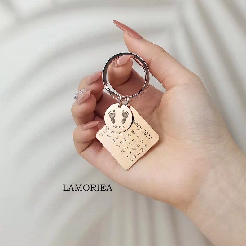May include: Gold-toned keychain with a square charm featuring a calendar for January 2021 and a round charm with a footprint design and the name "Emily".