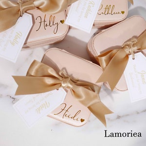 May include: Several blush-pink, personalized cosmetic cases with gold satin bows and tags. Each case is labeled with a name in gold script, such as "Holly," "Heidi," and "Kathleen." The tags read "Thank you for being my Bridesmaid."