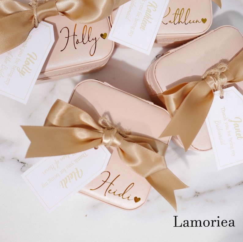 May include: Several blush-pink, personalized cosmetic cases with gold satin bows and tags. Each case is labeled with a name in gold script, such as "Holly," "Heidi," and "Kathleen." The tags read "Thank you for being my Bridesmaid."