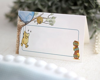 May include: A Winnie the Pooh themed place card with a blue balloon, a honey pot, and Winnie the Pooh holding onto a tree branch. The place card has a blank space for a name.