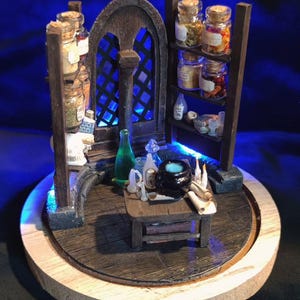 Può includere: Scena in miniatura di un'apoteca di streghe con una struttura in legno, scaffali e un tavolino. La scena comprende barattoli di vetro con coperchi in sughero, bottiglie, un calderone e pergamene. Lo sfondo ha una luce blu e la base è un cerchio di legno.