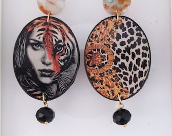 May include: A pair of oval drop earrings featuring a marbled circular stud. One earring displays a human face merged with a tiger, while the other shows a gold baroque pattern and leopard print. Each earring has a dangling black faceted bead.