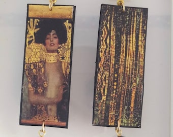 May include: A pair of gold and black earrings featuring a rectangular image of a woman with a gold background. The earrings have a black crystal dangle.