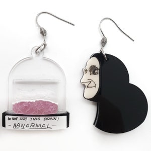 May include: A pair of quirky earrings. One earring features a clear dome with a pink brain inside, and the text "DO NOT USE THIS BRAIN! - ABNORMAL". The other earring is a black silhouette of a face with a white face and a grimacing expression.