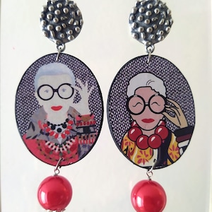 Puede incluir: Un par de pendientes de plata y rojo con un retrato de una mujer que lleva gafas y un collar colorido. Los pendientes son de forma ovalada y tienen una parte superior de plata texturizada.