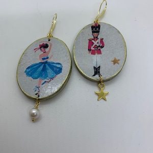 Può includere: Un paio di orecchini ovali con finitura dorata. Un orecchino presenta una ballerina con un tutù blu e l'altro un soldato giocattolo. Entrambi gli orecchini hanno un ciondolo a forma di stella dorata che pende dalla parte inferiore.